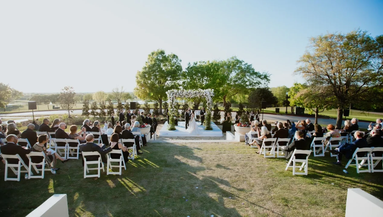 Outdoor wedding ceremony with guests seated on white chairs, facing a couple standing under a floral arch on a sunny day, surrounded by trees.