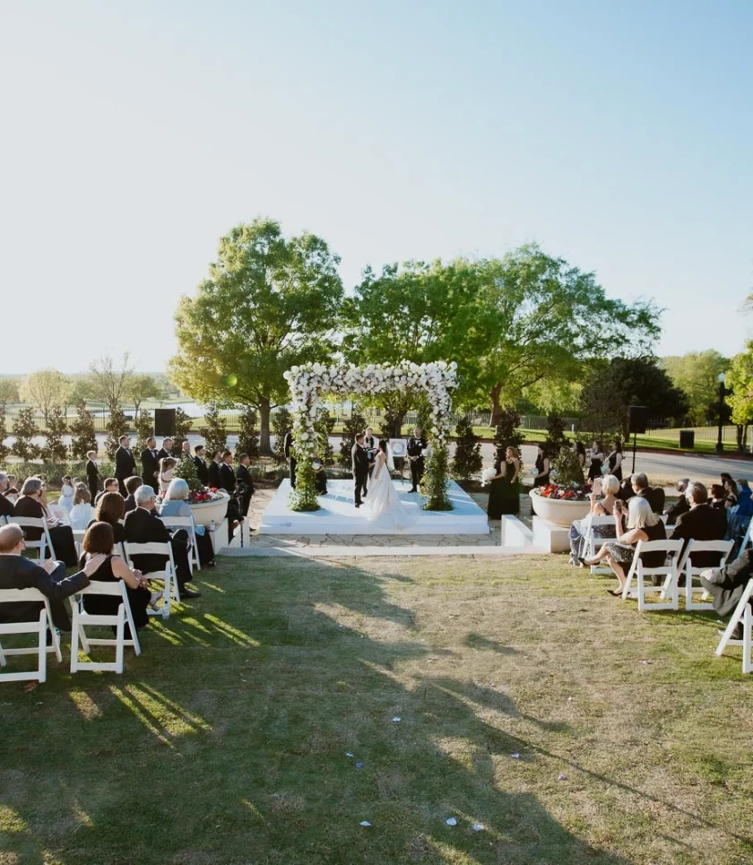 Outdoor wedding ceremony with guests seated on white chairs, facing a couple standing under a floral arch on a sunny day, surrounded by trees.