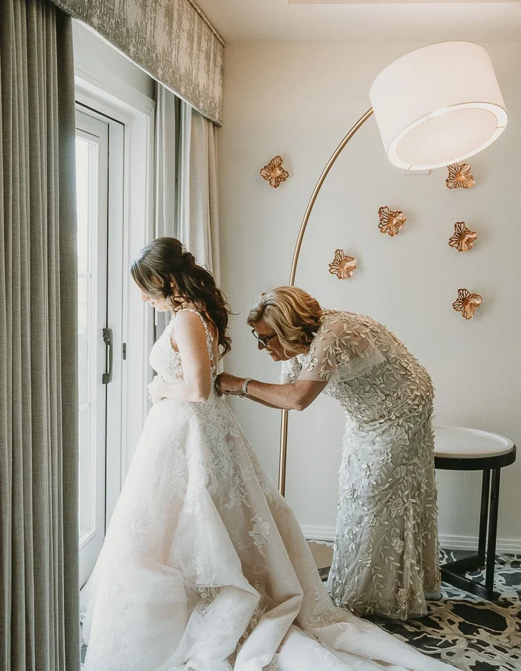 A woman helps a bride in a lace wedding dress with her gown near a window in a softly lit room decorated with gold butterfly wall art.
