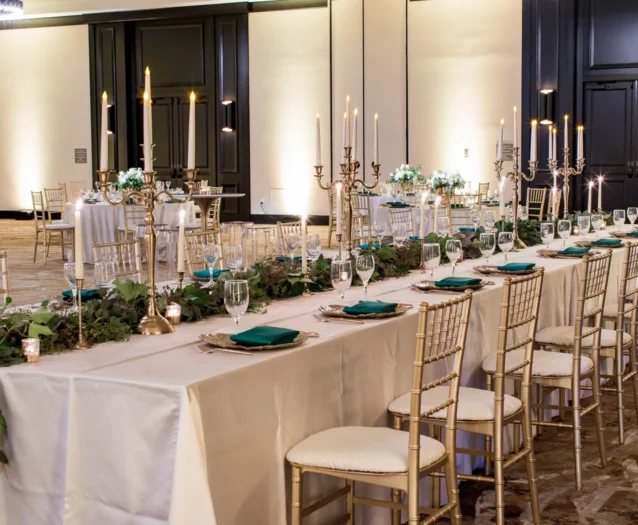 Elegant banquet hall with long tables set with white linens, gold chairs, green napkins, candles, and greenery centerpieces for a formal event.