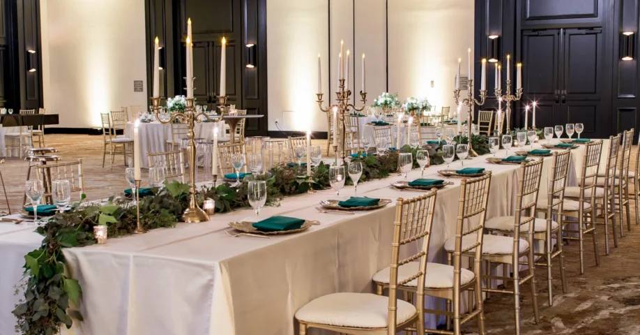 Elegant banquet hall with long tables set with white linens, gold chairs, green napkins, candles, and greenery centerpieces for a formal event.