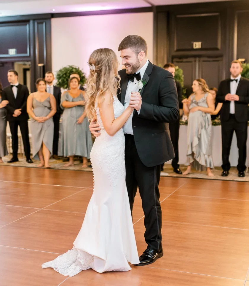 A bride and groom share their first dance as wedding guests in formal attire watch and smile in a spacious, elegant reception hall.