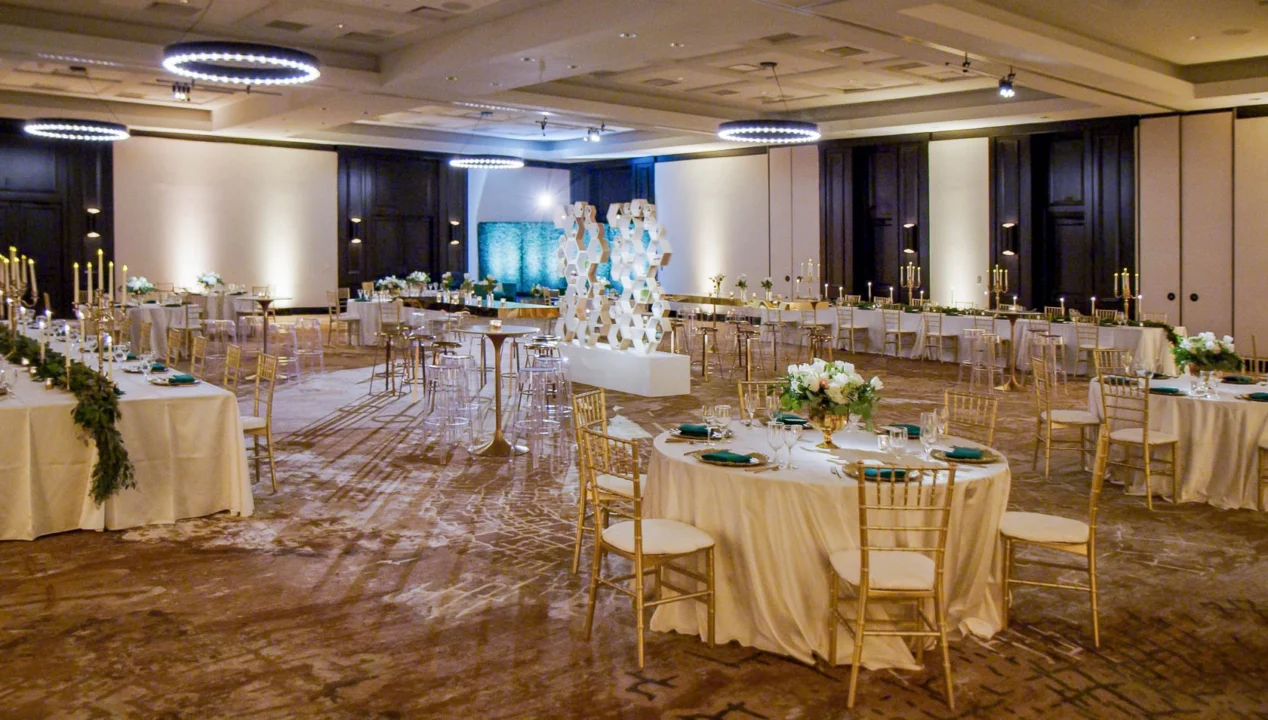Elegant wedding reception hall with round and rectangular tables, white tablecloths, gold chairs, floral centerpieces, and modern decor.