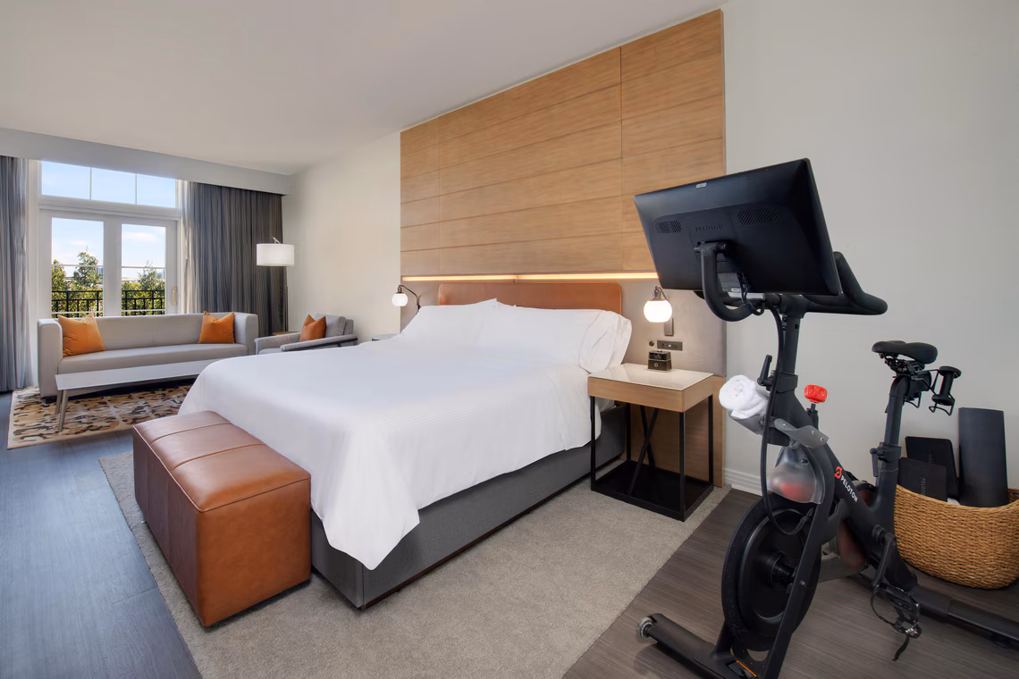 Modern hotel room with a king bed, exercise bike, sofa, large window, and neutral decor featuring wood and leather accents.