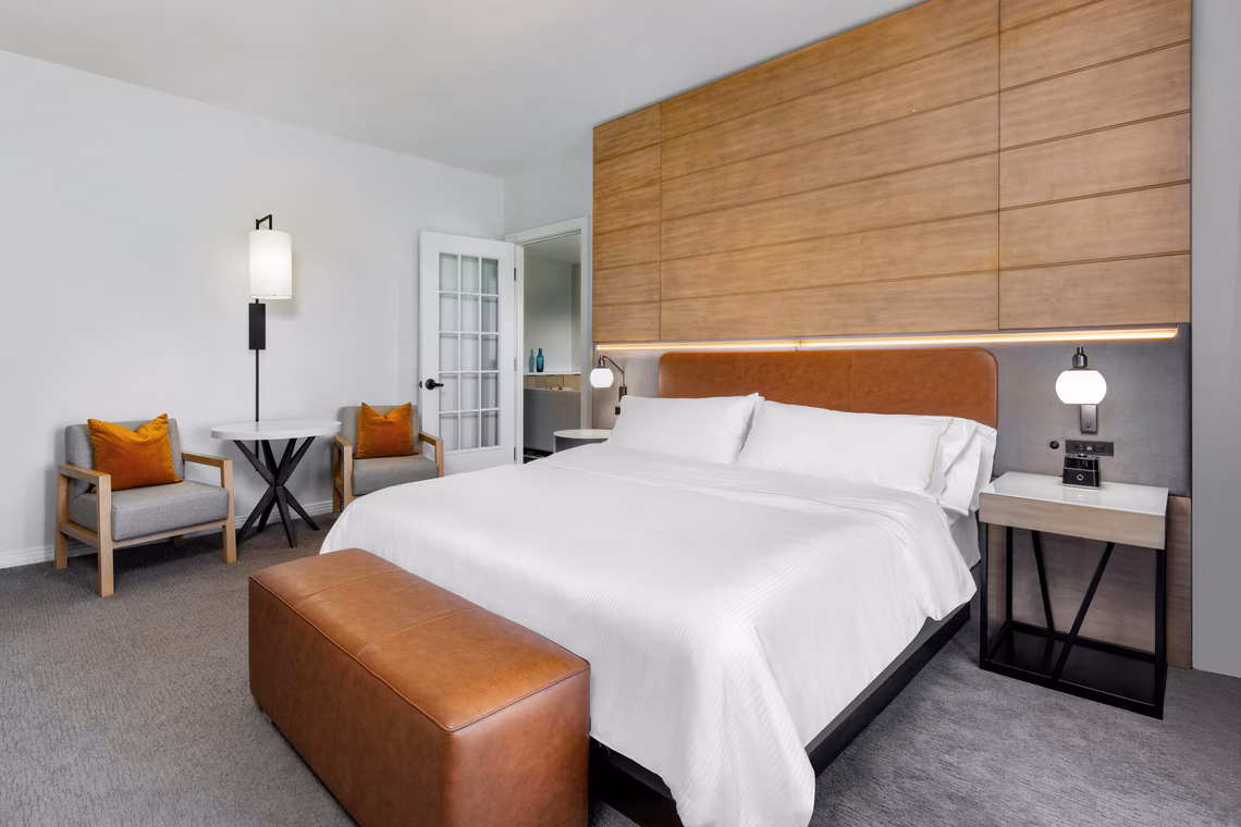 Modern hotel room with a king bed, two armchairs, side tables, a leather bench, and wooden accent wall in a neutral color palette.