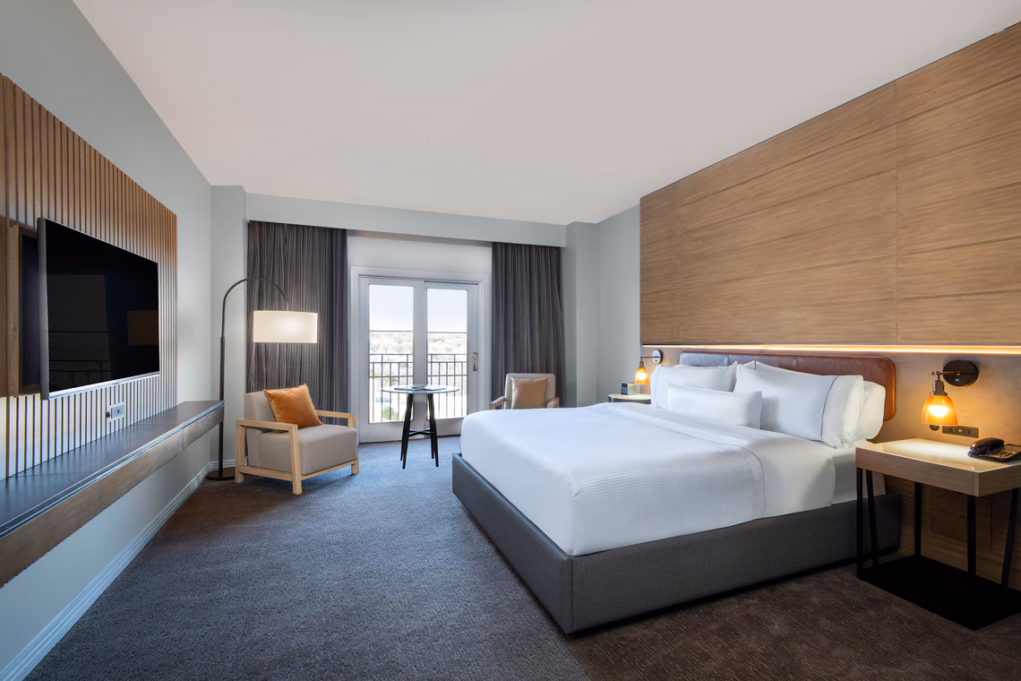 Modern hotel room with a king bed, armchair, TV, desk, and large window with balcony, decorated in neutral tones and wood accents.