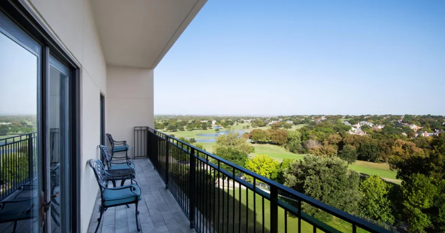 A spacious balcony with chairs overlooks a golf course, trees, and a distant neighborhood under a clear blue sky.