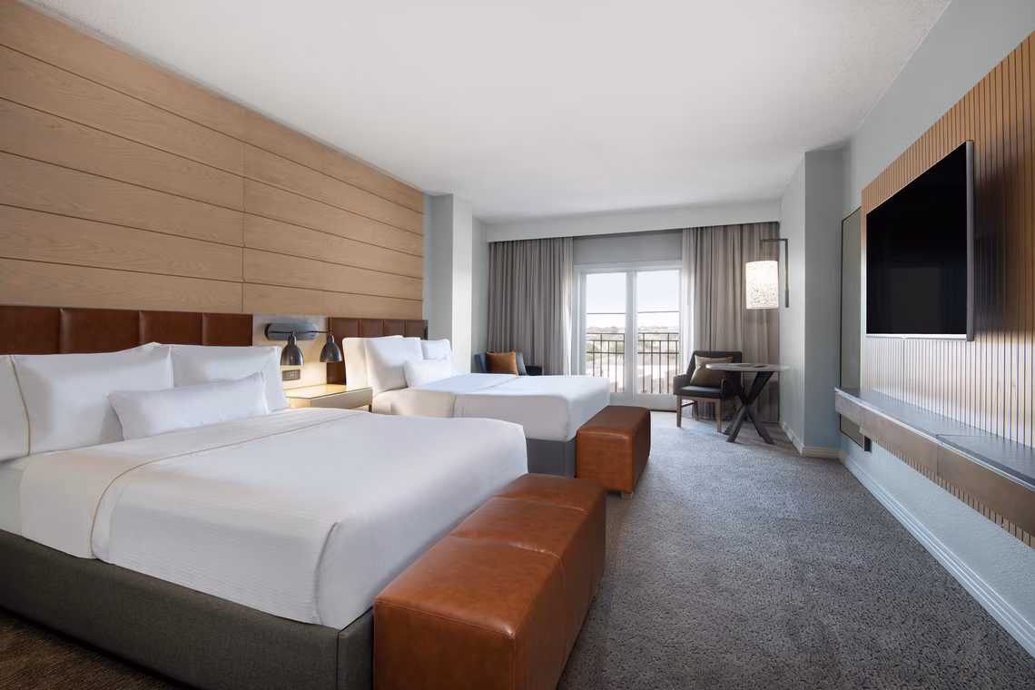 Modern hotel room with two large beds, a desk with a chair, a wall-mounted TV, and a window with curtains leading to a balcony.