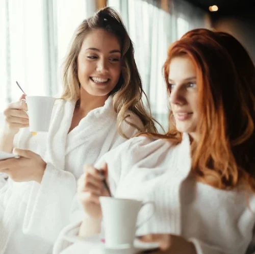 Two women in white robes smiling and relaxing with cups of coffee or tea, sitting together by a window with soft natural light.