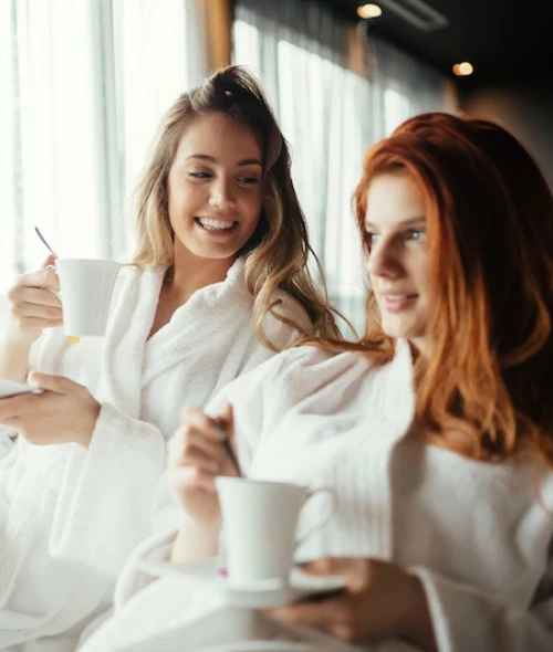 Two women in white robes smiling and relaxing with cups of coffee or tea, sitting together by a window with soft natural light.