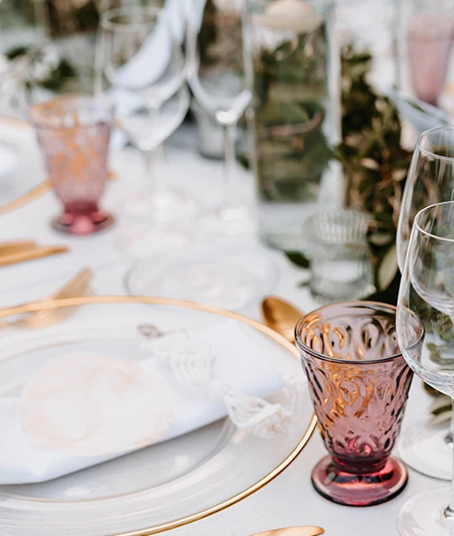 Elegant table setting with gold-rimmed plates, purple goblets, gold cutlery, and glassware on a white tablecloth with greenery decor.