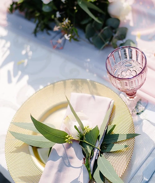 Elegant table setting with a gold charger plate, pale napkin, greenery, and a pink-tinted glass goblet on a white tablecloth.