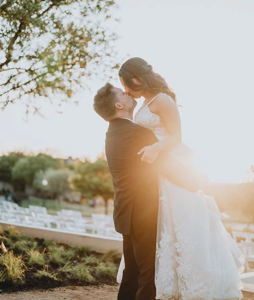 A groom lifts his bride for a sunset kiss outdoors at Golf Resort & SPA The Westin Stonebriar, framed by white chairs and lush greenery.
