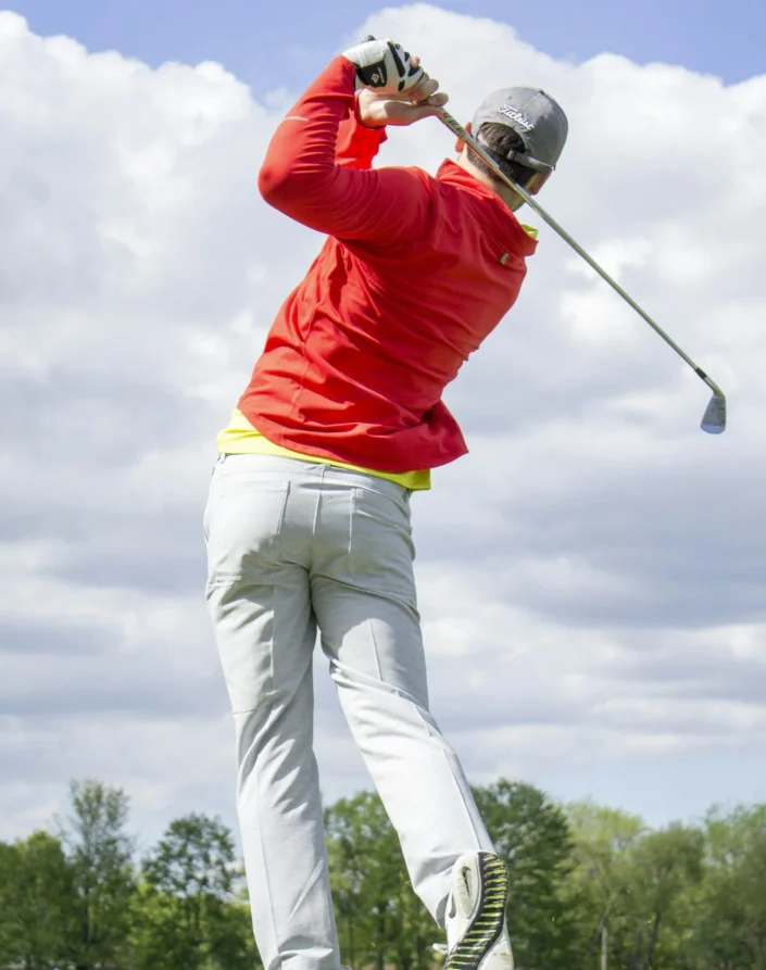 A golfer wearing a red jacket and gray pants swings a club on a grassy course under a partly cloudy sky.