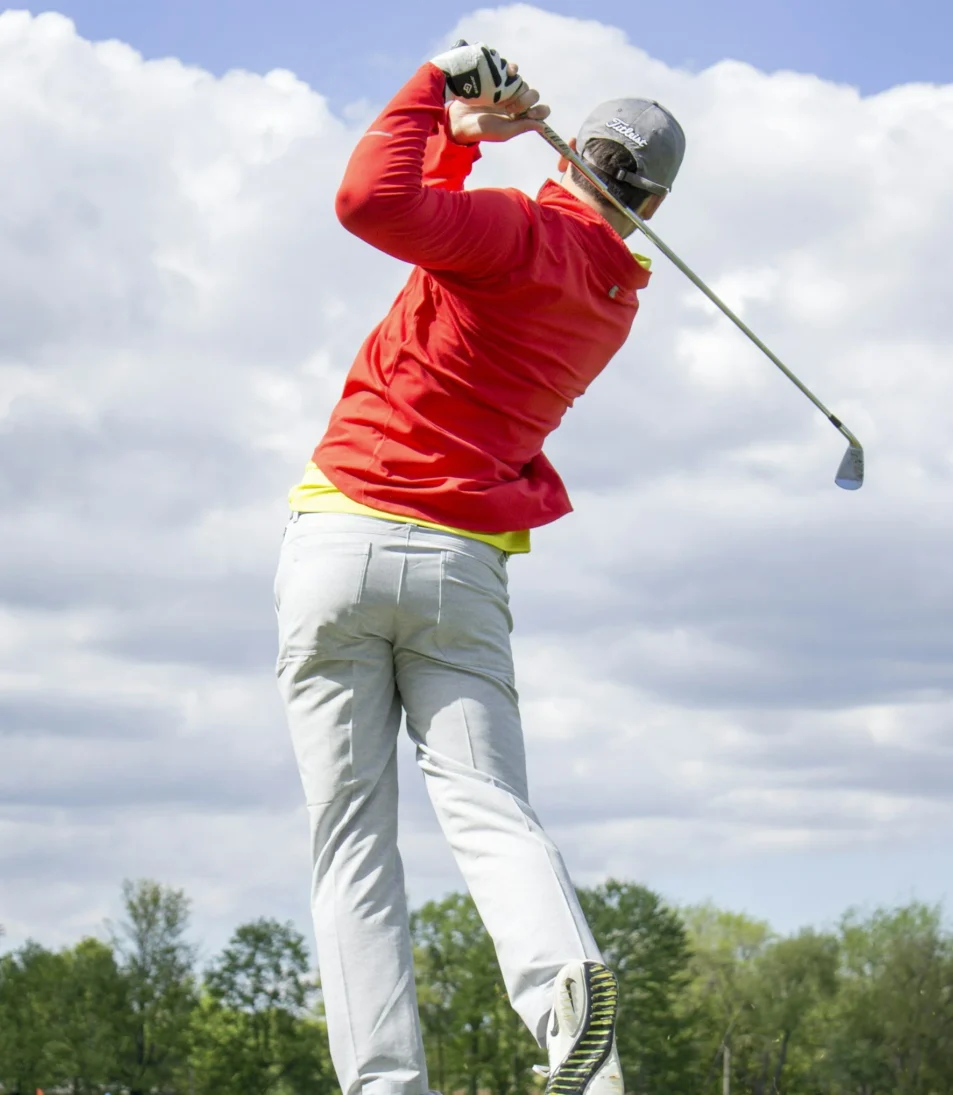 A golfer wearing a red jacket and gray pants swings a club on a grassy course under a partly cloudy sky.