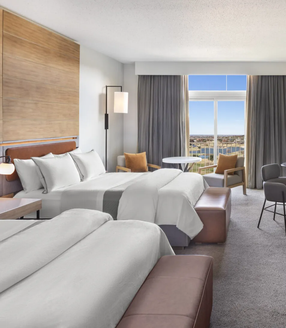 Modern hotel room with two double beds, a desk, seating area, large TV, and floor-to-ceiling window with city view.