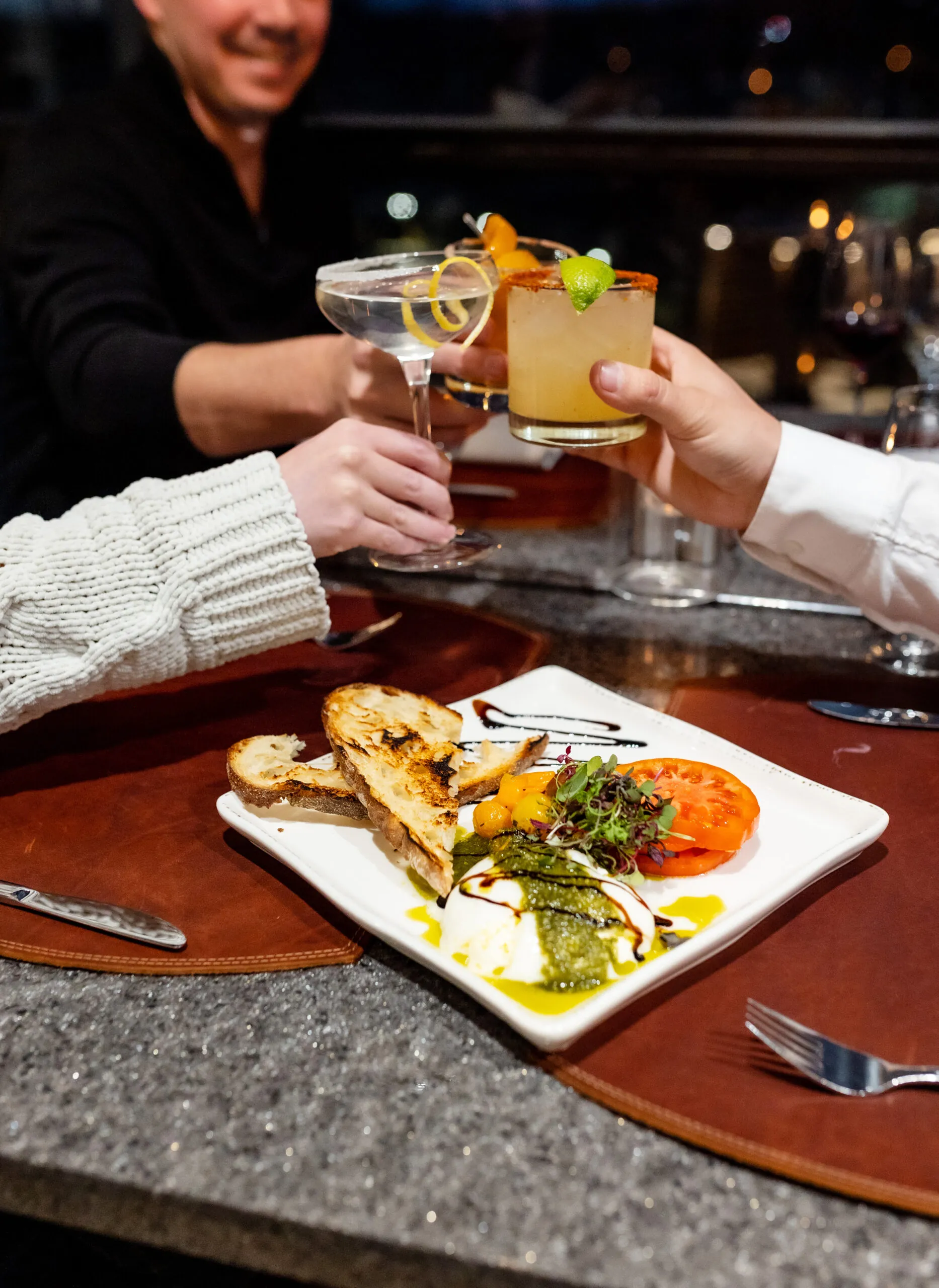 Three people toast with cocktails over a beautifully plated meal on a restaurant table, featuring grilled bread, tomato, cheese, and greens.