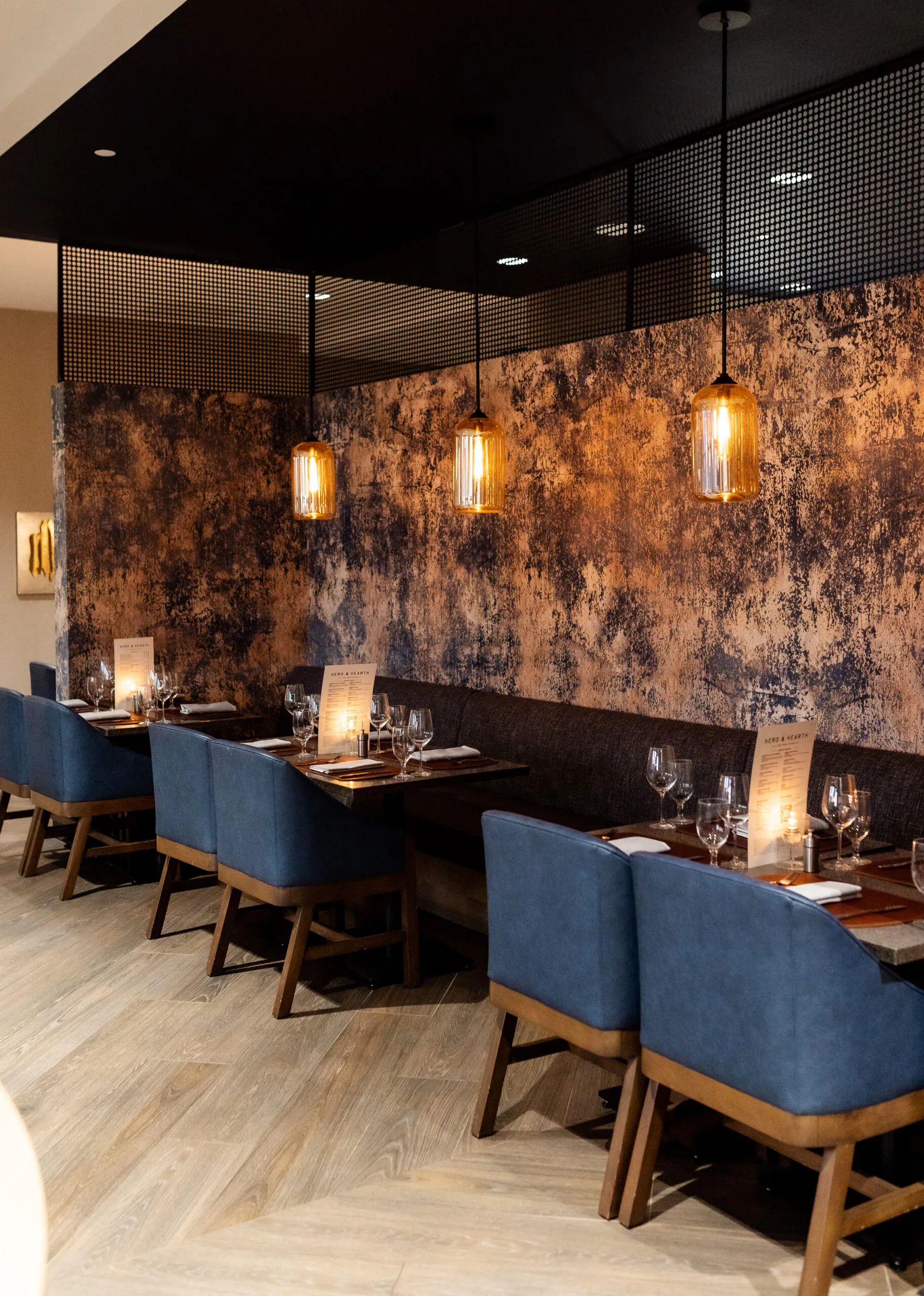 Elegant restaurant interior with blue chairs, wooden tables, hanging pendant lights, and textured wall decor, set for dining with glasses and menus.