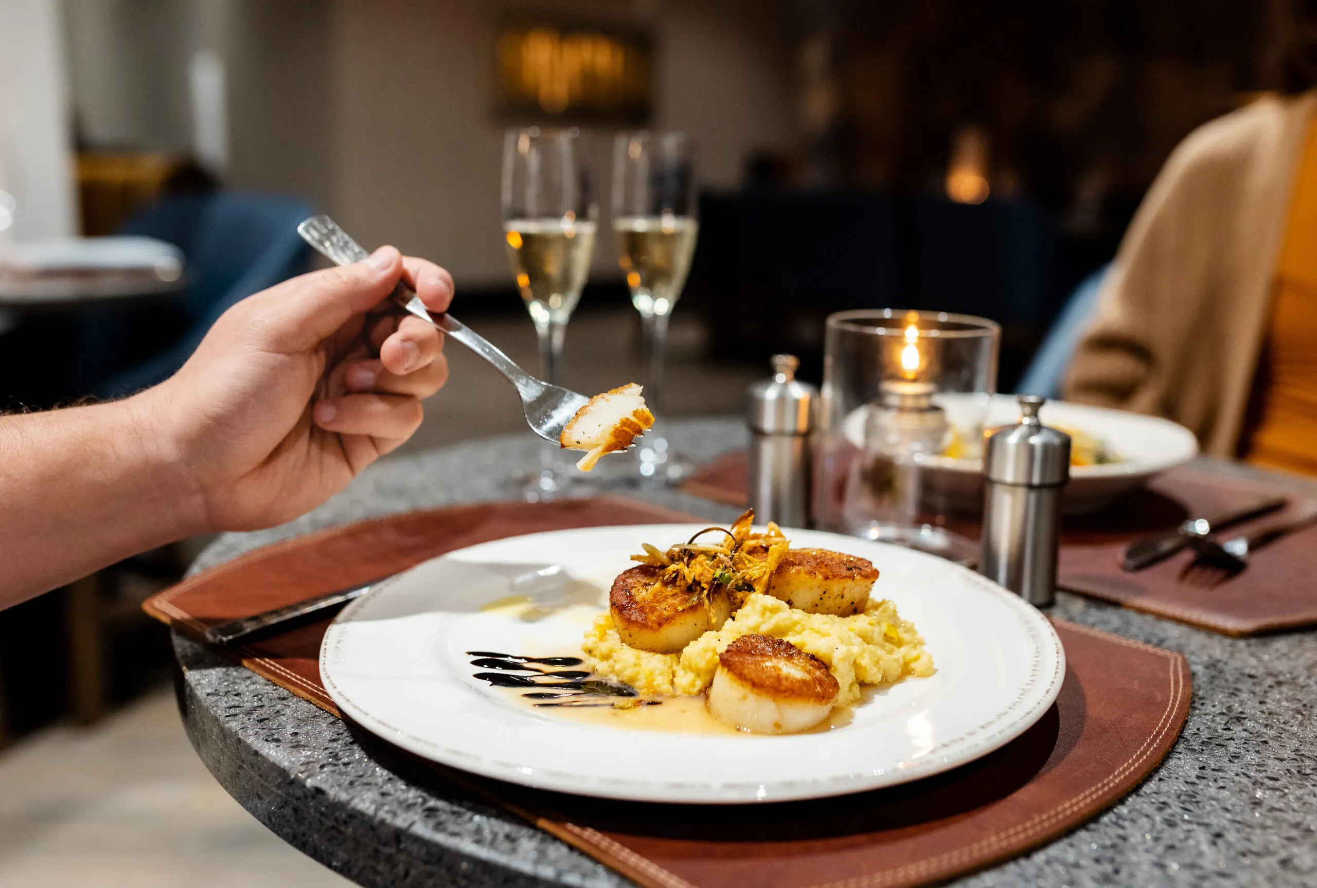 A hand holds a fork with a seared scallop over a plate of scallops and risotto, with wine glasses and candles on a restaurant table.
