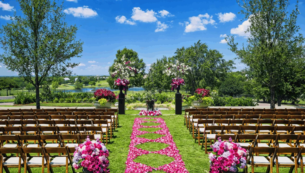 Outdoor wedding setup with rows of chairs, a pink flower petal aisle, floral arrangements, and trees under a bright blue sky.