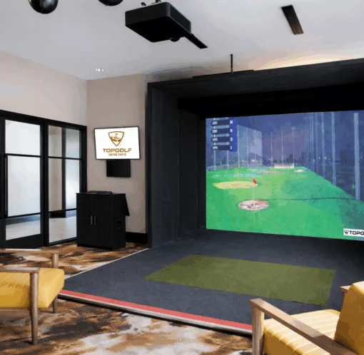 A modern indoor golf simulator room with a large screen, golf mat, two yellow chairs, and a Topgolf sign on the wall.