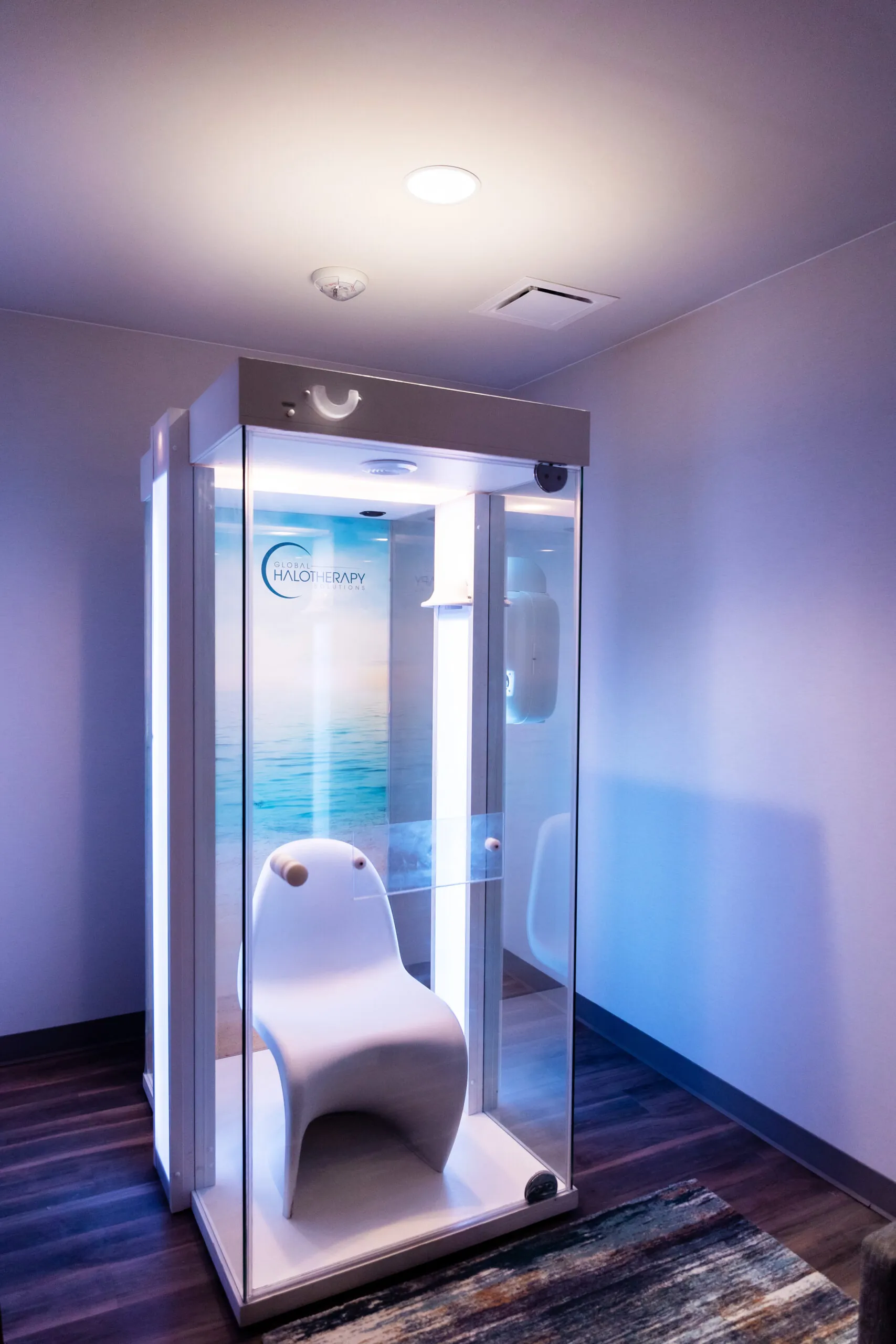 A modern halo therapy booth with a white chair inside, illuminated by soft lighting, sits in a corner of a room with wood flooring.