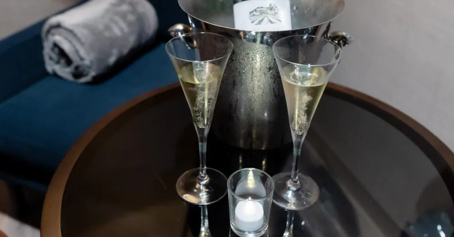 A chilled champagne bottle in an ice bucket with two filled glasses, a lit candle, and a Live It Well card on a round glass table.