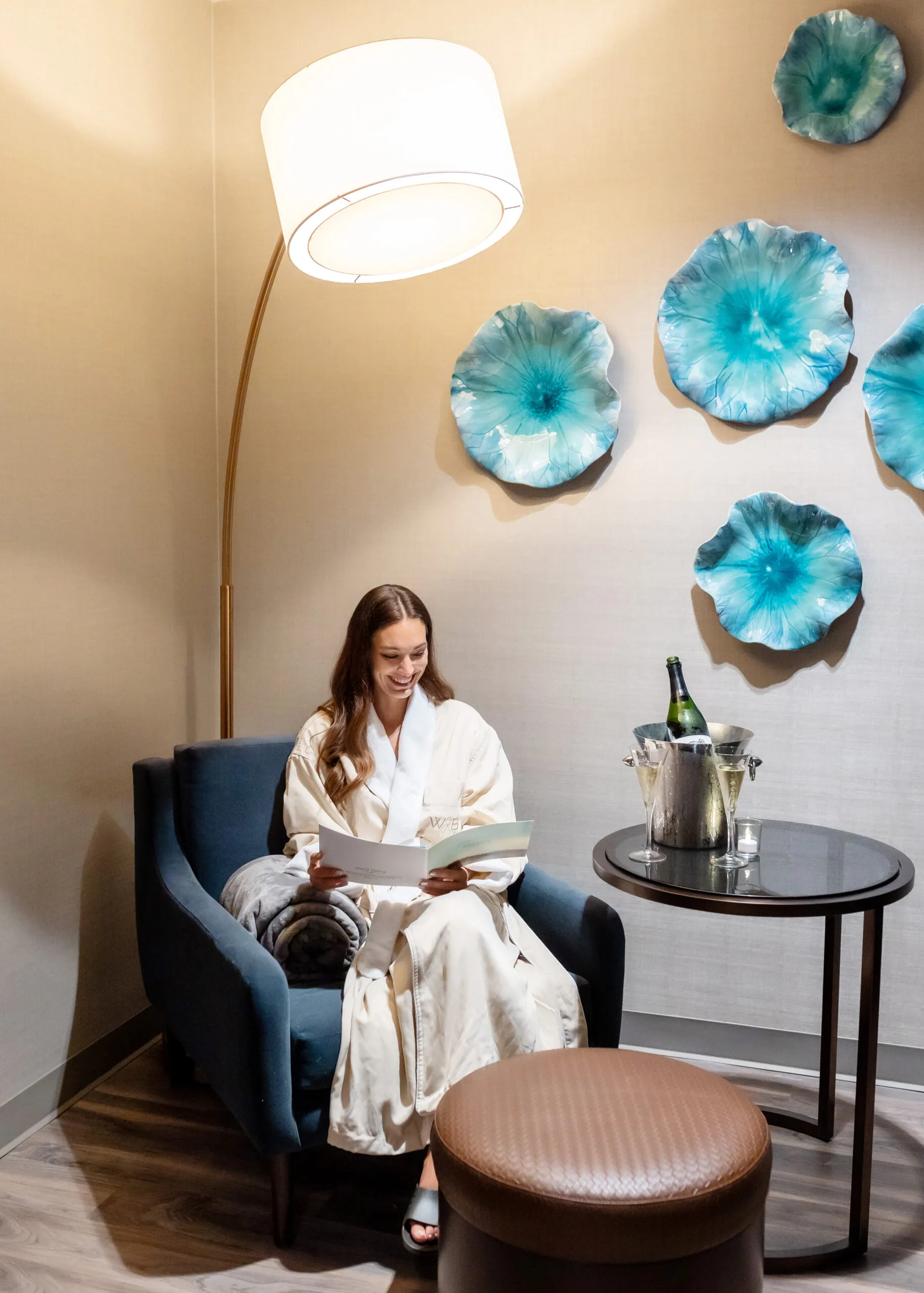A woman in a white robe sits in a cozy chair, reading, with champagne on a table and blue floral wall decor in a softly lit room.