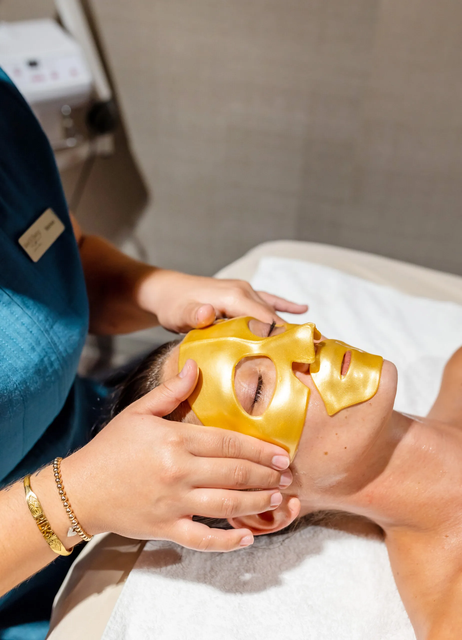 A person receives a facial treatment with a golden sheet mask while lying on a spa bed. A spa worker gently adjusts the mask on their face.