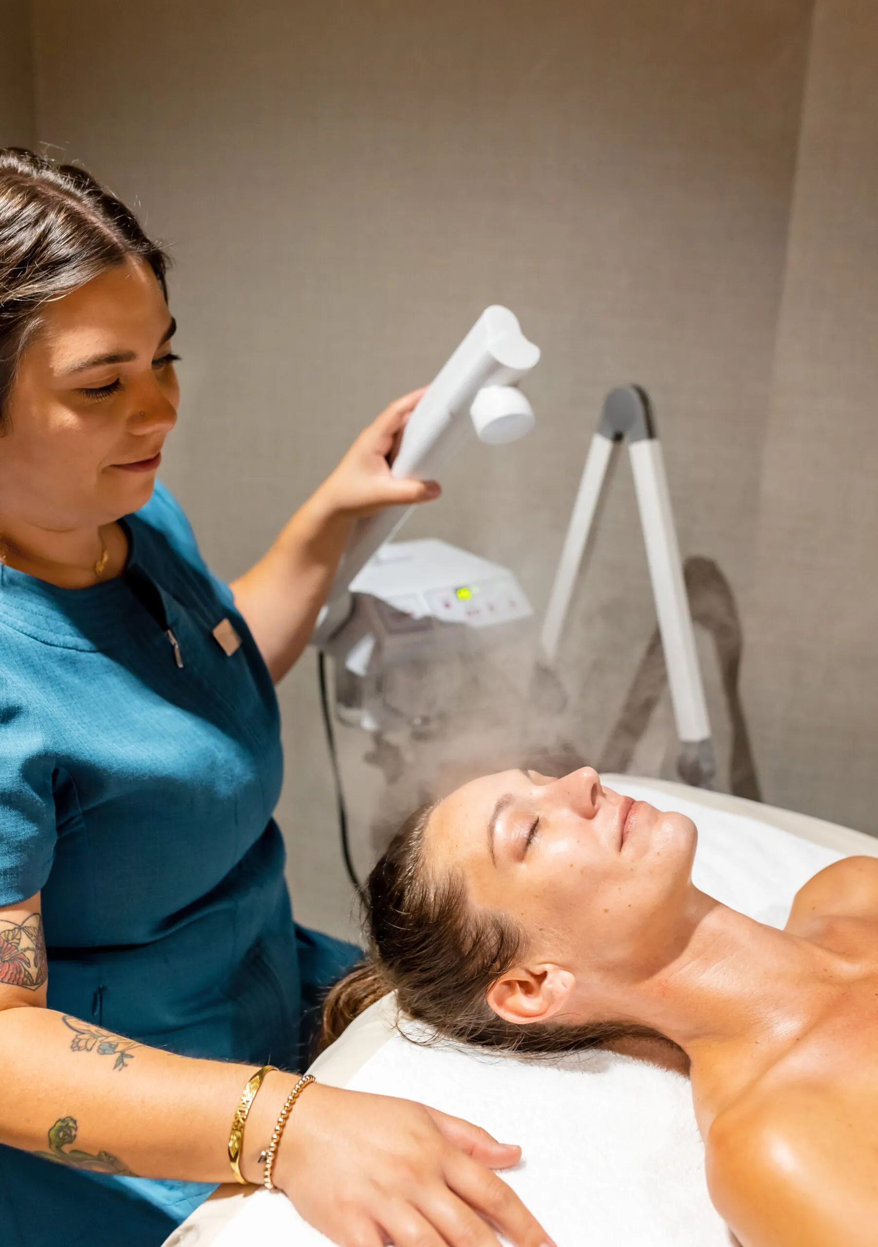 A woman receives a facial steam treatment from an esthetician in a spa setting, lying on a treatment bed with eyes closed.