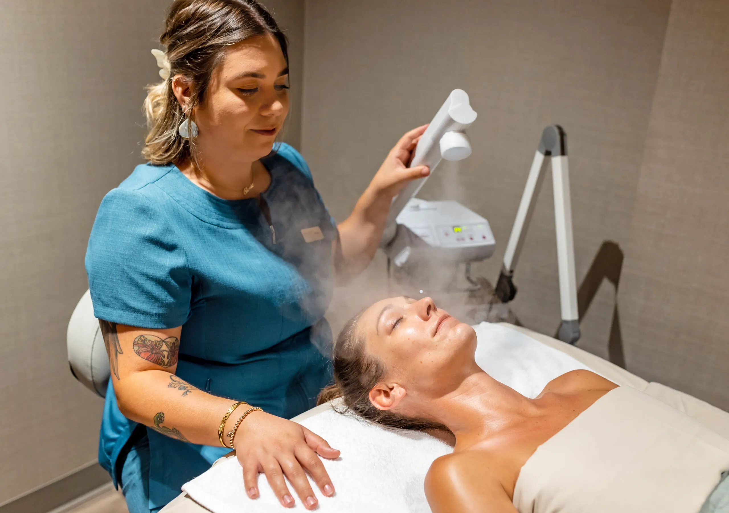 A spa therapist uses a facial steamer on a woman lying on a treatment table, both in a calm, relaxing spa room.
