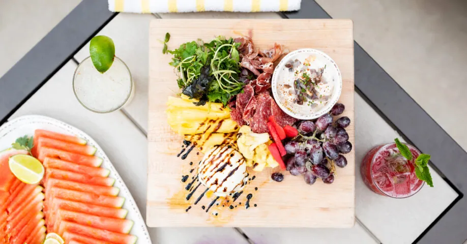 Charcuterie board with meats, cheese, grapes, microgreens, a drink with lime, a drink with mint, watermelon slices, and purple sunglasses on a table.
