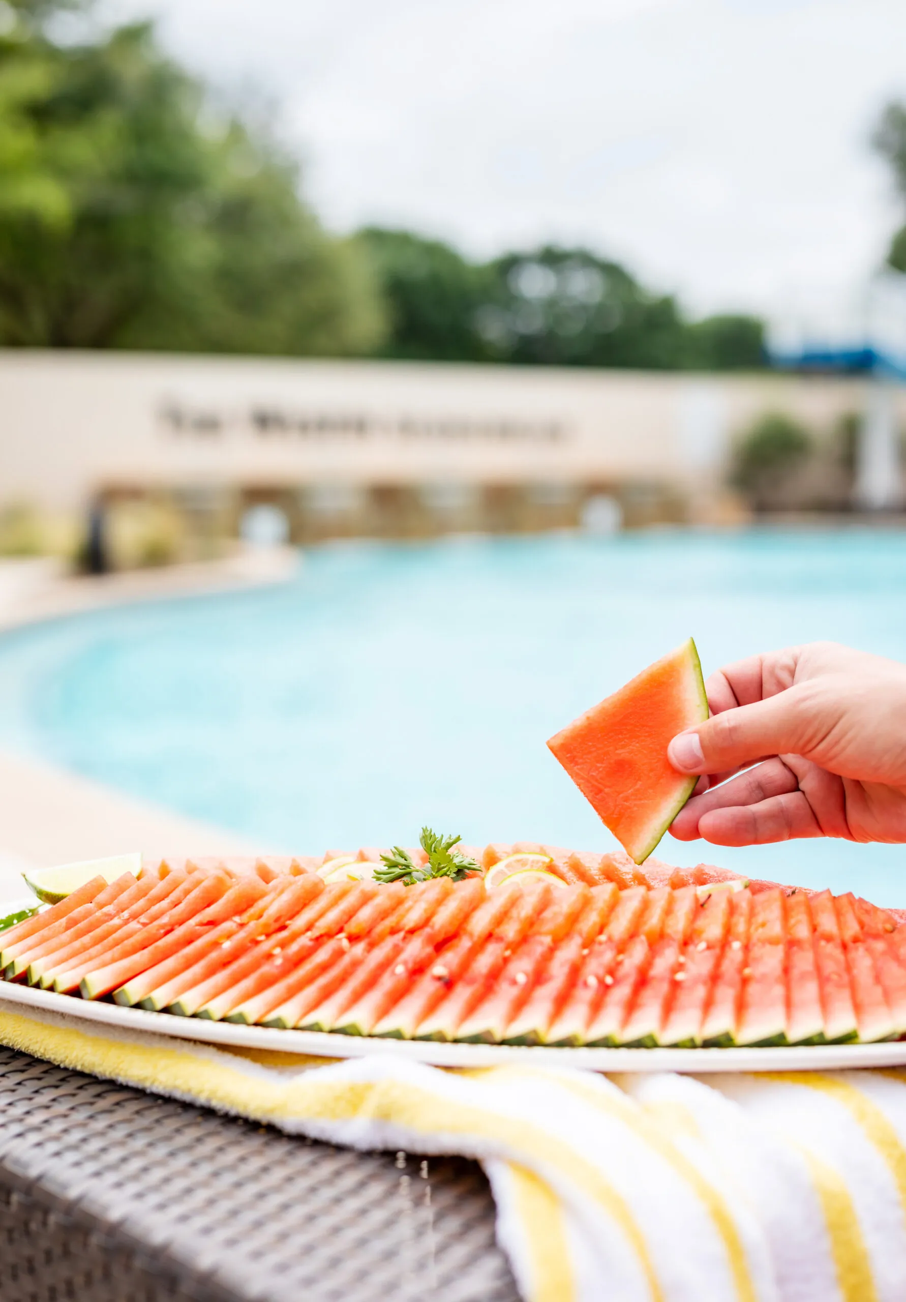 A hand holds a slice of watermelon by a pool, with a platter of neatly arranged watermelon slices on a towel nearby.