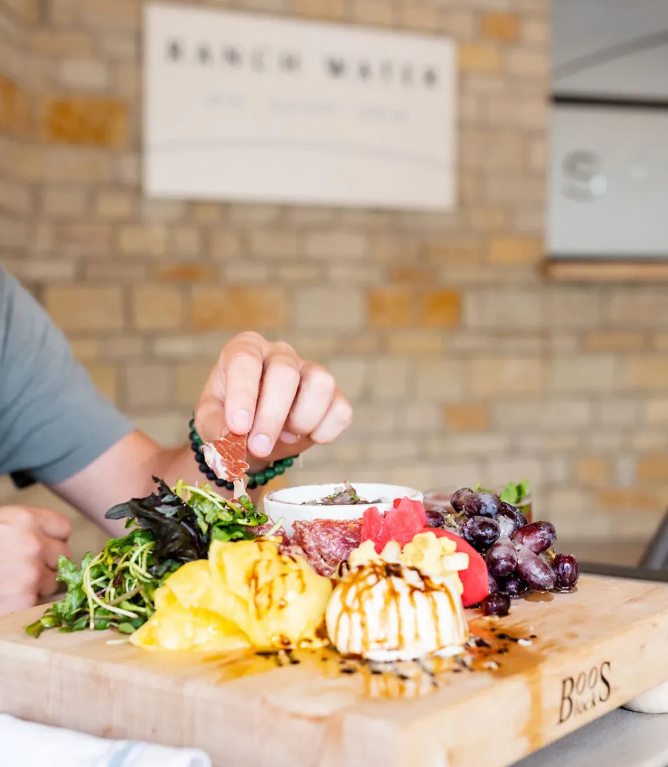 A hand reaches for food on a wooden charcuterie board with cheese, fruit, greens, and meat, set on a table in front of a brick wall.