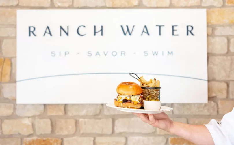 A hand holds a plate with a burger, fries in a metal basket, and a dipping sauce in front of a sign reading RANCH WATER SIP • SAVOR • SWIM.