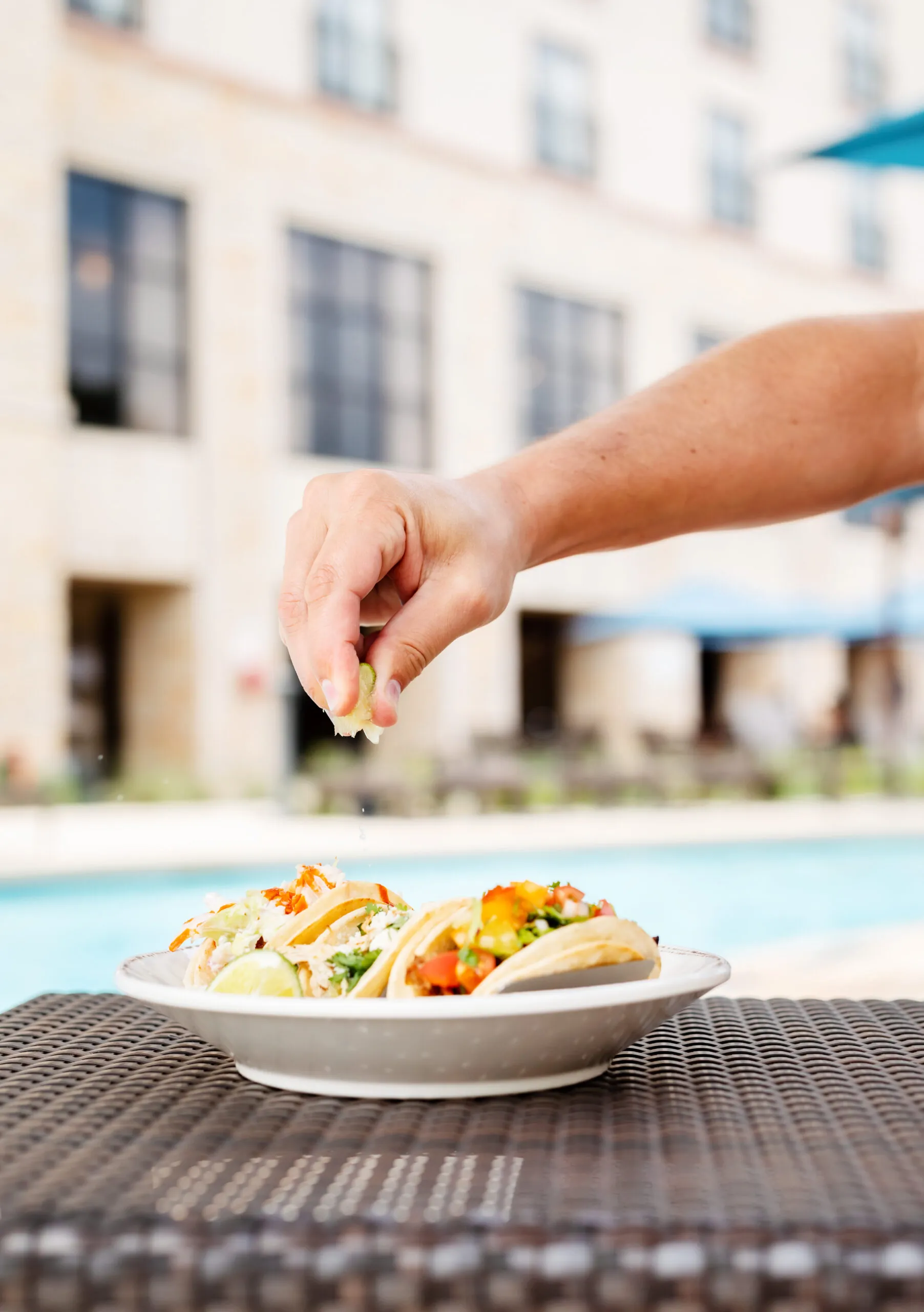 A hand squeezes a lime over a plate of tacos on an outdoor table near a pool, with a building in the blurred background.
