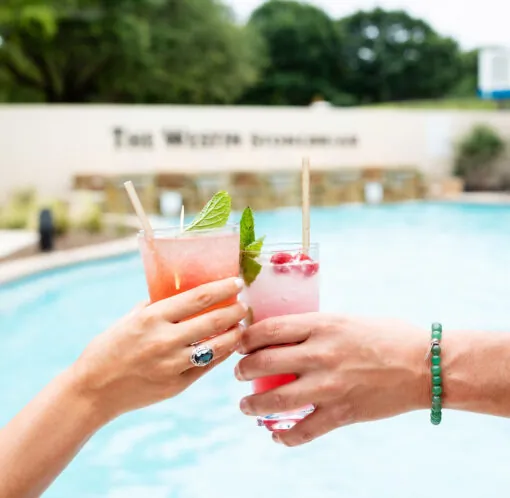 Two hands clink colorful cocktails with straws by a swimming pool, with trees and a stone wall in the background.