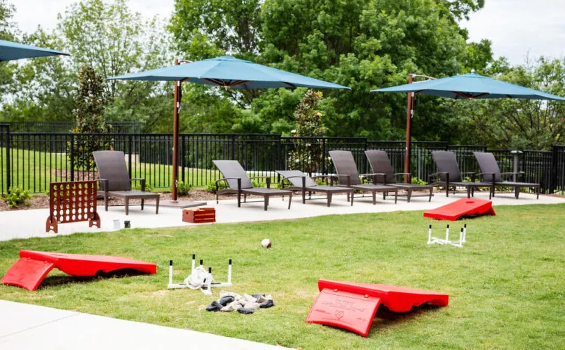Lawn with red cornhole boards, outdoor lounge chairs, large blue umbrellas, and yard games, surrounded by green trees and a black fence.