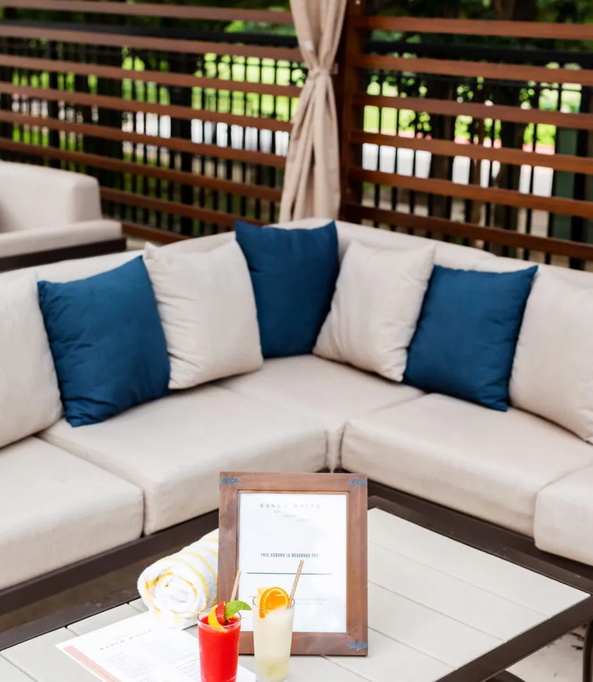 Outdoor patio seating with beige cushions, blue pillows, a table holding two cocktails, a menu, and a rolled towel, with greenery in the background.