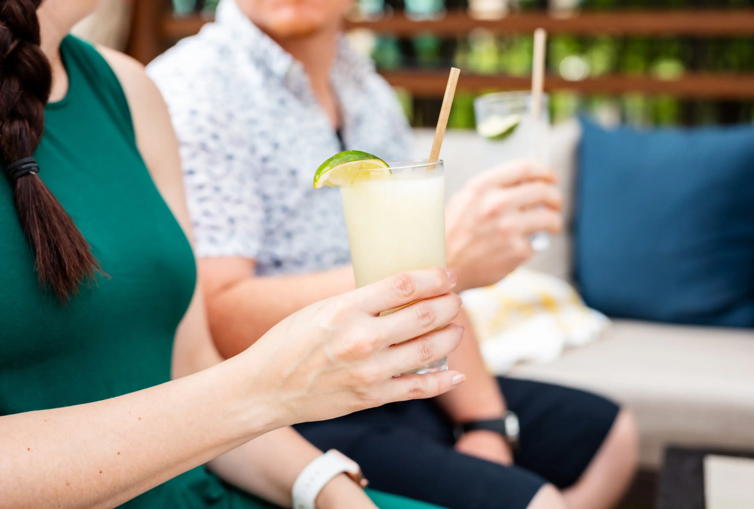 Two people sit outdoors holding cocktails with lime garnishes, one wearing a green dress, the other a patterned shirt and shorts, enjoying a relaxed moment.