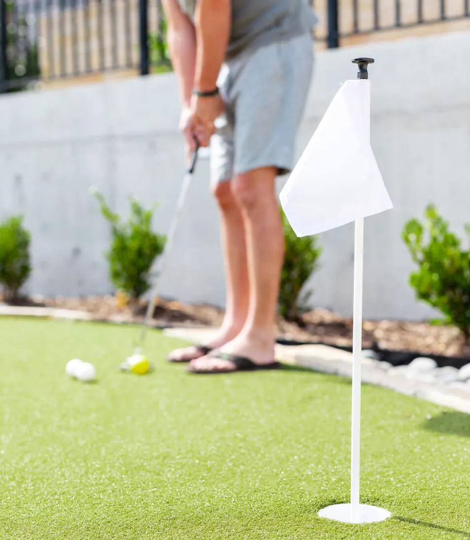 A person in shorts putts a golf ball on a green, with a small white flag in the foreground and bushes in the background.