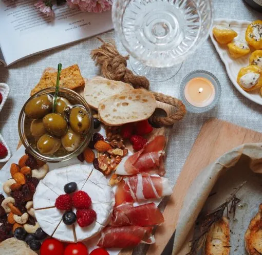 A charcuterie board with bread, olives, nuts, cheese topped with berries, tomatoes, crackers, prosciutto, and a glass of wine on a table.