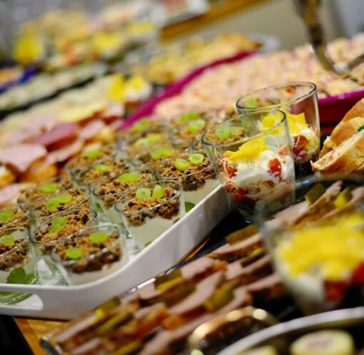 A close-up of a buffet table with assorted appetizers, including layered parfaits, sliced bread, meats, cheeses, and colorful garnished dishes.