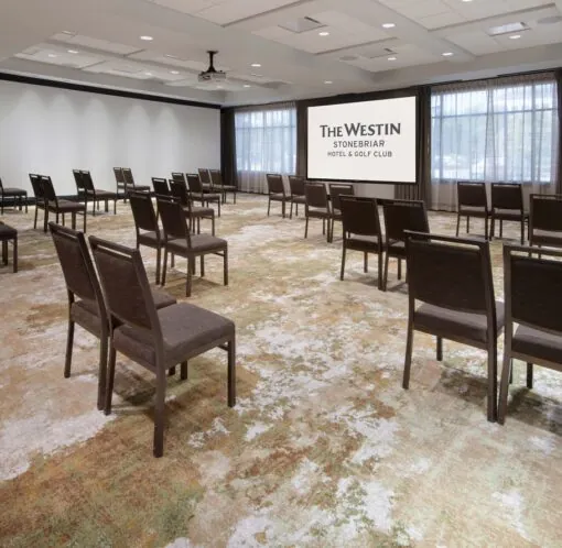 A conference room with spaced-out chairs facing a screen that reads The Westin Stonebriar Hotel & Golf Club, with large windows and neutral decor.