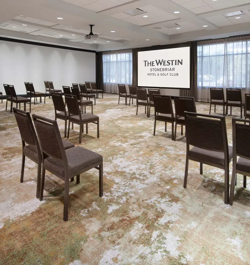 A conference room with spaced-out chairs facing a screen that reads The Westin Stonebriar Hotel & Golf Club, with large windows and neutral decor.