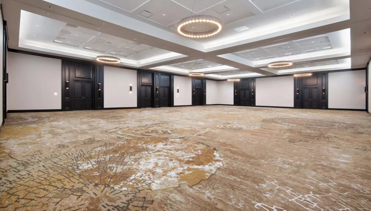 Spacious, empty ballroom with patterned carpet, modern chandeliers, white ceiling panels, and dark wood doors along the walls.