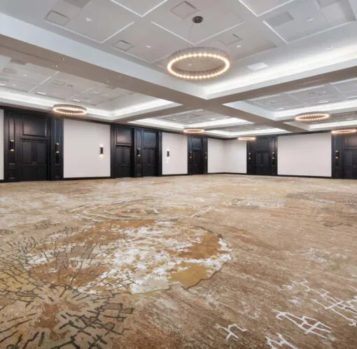 Spacious, empty ballroom with patterned carpet, modern chandeliers, white ceiling panels, and dark wood doors along the walls.
