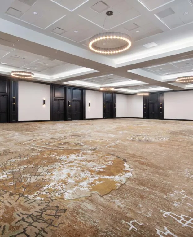 Spacious, empty ballroom with patterned carpet, modern chandeliers, white ceiling panels, and dark wood doors along the walls.