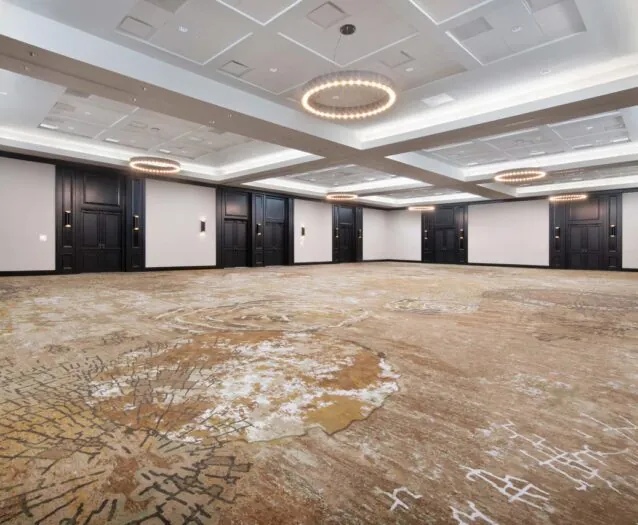Spacious, empty ballroom with patterned carpet, modern chandeliers, white ceiling panels, and dark wood doors along the walls.
