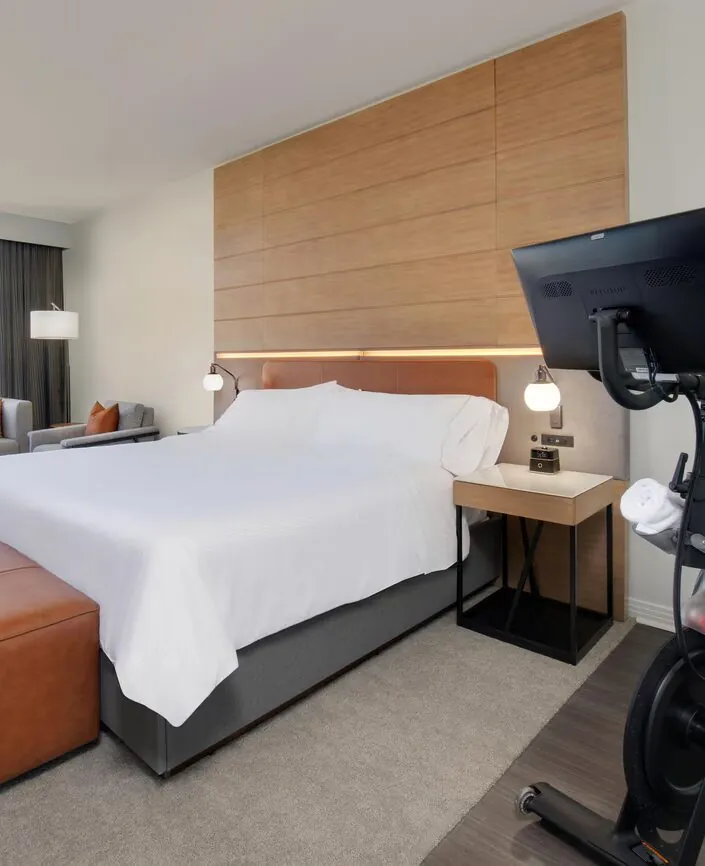 Modern hotel room with a neatly made bed, exercise bike, sofa, large window, and contemporary decor.